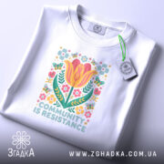 Футболка єдність і сила, біла футболка, складена на поверхні, з принтом квітів і написом 'COMMUNITY IS RESISTANCE', фон світло-фіолетовий, видно етикетку з логотипом.