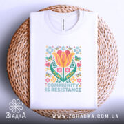 Футболка єдність і сила, біла футболка, складена на плетеній кошику, з принтом квітів і написом 'COMMUNITY IS RESISTANCE', фон світлий, видно етикетку з логотипом.