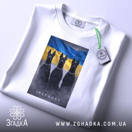 Футболка інстинкт білого кольору, розкладена на поверхні. На ній принт із трьома чорними собаками на фоні жовто-блакитного прапора. Видно етикетку з логотипом, що підвішена на зеленій стрічці. Фон нейтральний, що дозволяє акцентувати увагу на футболці.