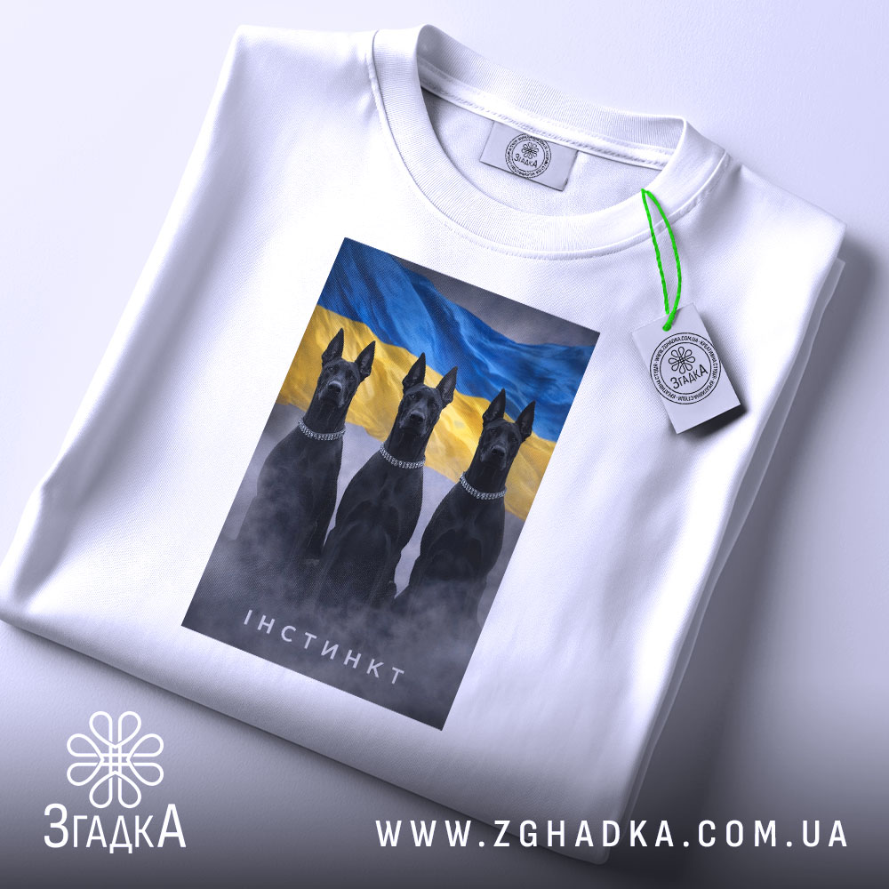 Футболка інстинкт білого кольору, розкладена на поверхні. На ній принт із трьома чорними собаками на фоні жовто-блакитного прапора. Видно етикетку з логотипом, що підвішена на зеленій стрічці. Фон нейтральний, що дозволяє акцентувати увагу на футболці.