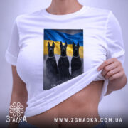 Футболка інстинкт білого кольору, розкладена на поверхні. На ній принт із трьома чорними собаками на фоні жовто-блакитного прапора. Видно етикетку з логотипом, що підвішена на зеленій стрічці. Фон нейтральний, що дозволяє акцентувати увагу на футболці.