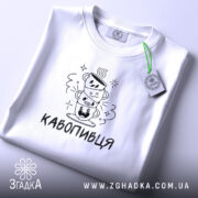 Футболка кавопивця білого кольору, складена на поверхні, ракурс зверху, фон світло-фіолетовий, принт з трьома чашками кави, одна з чашок з усмішкою, інша з закритими очима, текст 'КАВОПИВЕЦЬ' під принтом, на комірці етикетка.