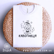 Футболка кавопивця білого кольору, складена на плетеній кошику, ракурс зверху, фон світло-фіолетовий, принт з трьома чашками кави, одна з чашок з усмішкою, інша з закритими очима, текст 'КАВОПИВЕЦЬ' під принтом.