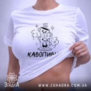 Футболка кавопивця білого кольору, складена на поверхні, ракурс зверху, фон світло-фіолетовий, принт з трьома чашками кави, одна з чашок з усмішкою, інша з закритими очима, текст 'КАВОПИВЕЦЬ' під принтом, на комірці етикетка.