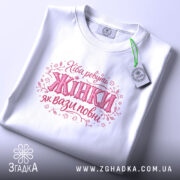 Футболка хіба ревуть жінки, біла, зображена на плоскій поверхні, фон світло-сірий, принт з написом 'Хіба ревуть Жінки як вази повні' в рожевих тонах, з етикеткою зеленого кольору.