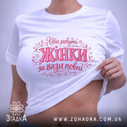 Футболка хіба ревуть жінки, біла, зображена на плоскій поверхні, фон світло-сірий, принт з написом 'Хіба ревуть Жінки як вази повні' в рожевих тонах, з етикеткою зеленого кольору.