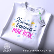 Футболка хто має терпіння, біла, складена на поверхні, фон світло-сірий, принт з текстом 'Хто має терпіння – має всіх' у кольорах синьому, рожевому та жовтому, з зірочками, на етикетці зелений ярлик.