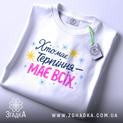 Футболка хто має терпіння, біла, складена на поверхні, фон світло-сірий, принт з текстом 'Хто має терпіння – має всіх' у кольорах синьому, рожевому та жовтому, з зірочками, на етикетці зелений ярлик.
