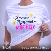 Футболка хто має терпіння, біла, складена на поверхні, фон світло-сірий, принт з текстом 'Хто має терпіння – має всіх' у кольорах синьому, рожевому та жовтому, з зірочками, на етикетці зелений ярлик.