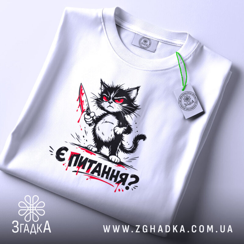 Футболка кіт є питання, біла футболка, складена на поверхні, принт з котом з ножем, фон світлий, присутня етикетка з зеленим шнурком.