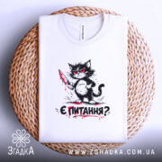 Футболка кіт є питання, біла футболка, складена на плетеній кошику, принт з котом з ножем, фон світлий, деталі принта яскраві, видно етикетку.