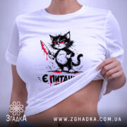 Футболка кіт є питання, біла футболка, складена на поверхні, принт з котом з ножем, фон світлий, присутня етикетка з зеленим шнурком.