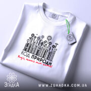 Футболка код природи екостиль, біла, на столі, з етикеткою, фон світло-сірий, принт з рослинними елементами та написом 'КОД ПРИРОДИ! Радій, кожний мить жити!' у чорному та червоному кольорах.