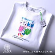 Футболка кохання це біла футболка, складена на поверхні з м'яким освітленням. Принт на футболці показує двох анімованих персонажів, кішку і собаку, з написом 'Кохання це...' над ними. Футболка має круглий виріз і виглядає акуратно.