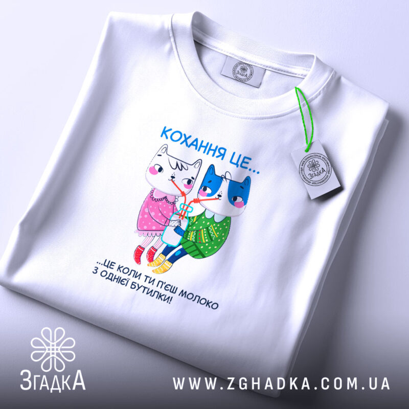 Футболка кохання це біла футболка, складена на поверхні з м'яким освітленням. Принт на футболці показує двох анімованих персонажів, кішку і собаку, з написом 'Кохання це...' над ними. Футболка має круглий виріз і виглядає акуратно.