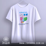 Футболка кохання це біла футболка, підвішена на вішалці з нейтральним фоном. Принт на футболці містить двох анімованих персонажів, кішку і собаку, з написом 'Кохання це...' під ними. Футболка має короткі рукави і простий дизайн.