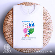 Футболка кохання це біла футболка, складена на плетеній кошику з нейтральним фоном. Принт на футболці зображує двох анімованих персонажів, кішку і собаку, з написом 'Кохання це...' під ними. Футболка має круглий виріз і виглядає стильно.