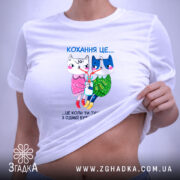 Футболка кохання це біла футболка, складена на поверхні з м'яким освітленням. Принт на футболці показує двох анімованих персонажів, кішку і собаку, з написом 'Кохання це...' над ними. Футболка має круглий виріз і виглядає акуратно.