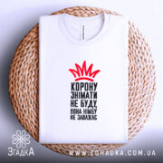 Футболка корону знімати не буду, біла футболка на плоскому фоні, зображена зверху на плетеній основі, принт червоного кольору у вигляді корони та чорного тексту, видна етикетка.