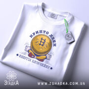 Футболка крипто дзен, біла, на плоскій поверхні, з принтом у вигляді усміхненої монети з символом біткоїна, текстом 'крипто дзен' та 'енергія блокчейну', фон світло-сірий.