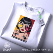 Футболка кумедний подарунок подрузі, біла, зображена на столі, фон світло-сірий, принт з дівчиною, що тримає два келихи з напоями, один жовтий, інший червоний, з текстом внизу, на футболці є етикетка.