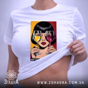 Футболка кумедний подарунок подрузі, біла, зображена на столі, фон світло-сірий, принт з дівчиною, що тримає два келихи з напоями, один жовтий, інший червоний, з текстом внизу, на футболці є етикетка.