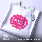 Футболка мама найкраща, біла, складена на столі, фон світло-фіолетовий, принт рожевий з написом 'НАЙКРАЩА МАМА У СВІТІ' у формі герба, з етикеткою.