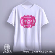 Футболка мама найкраща, біла, підвішена на вішалці, фон світло-сірий, принт рожевий з написом 'НАЙКРАЩА МАМА У СВІТІ' у формі герба.