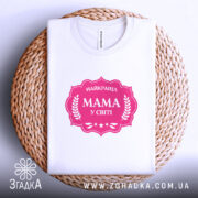 Футболка мама найкраща, біла, складена на плетеній кошику, фон білий, принт рожевий з написом 'НАЙКРАЩА МАМА У СВІТІ' у формі герба.