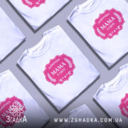 Футболка мама найкраща, біла, на моделі, фон світло-фіолетовий, принт рожевий з написом 'НАЙКРАЩА МАМА У СВІТІ' у формі герба.