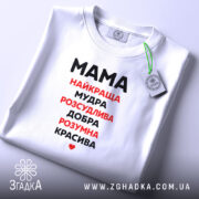 Футболка мама найкраща, біла, зображена на плоскій поверхні, фон світло-сірий, принт чорного та червоного кольору з написом 'Мама найкраща, мудра, розсудлива, добра, розумна, красива', з етикеткою на комірці.