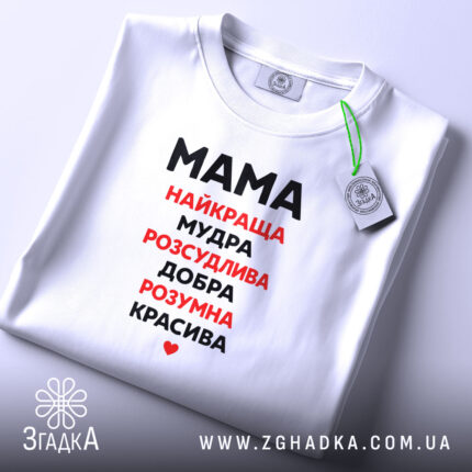 Футболка мама найкраща, біла, зображена на плоскій поверхні, фон світло-сірий, принт чорного та червоного кольору з написом 'Мама найкраща, мудра, розсудлива, добра, розумна, красива', з етикеткою на комірці.