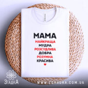 Футболка мама найкраща, біла, зображена на плоскій поверхні, фон світлий, футболка лежить на плетеній основі, принт чорного та червоного кольору з написом 'Мама найкраща, мудра, розсудлива, добра, розумна, красива'.