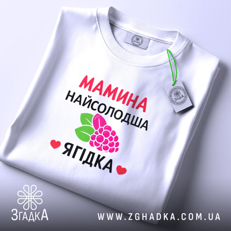 Футболка мамина найсолодша ягідка, біла, складена на поверхні, фон світло-сірий, принт з червоним текстом та малюнком ягід і листя, з етикеткою на комірці.