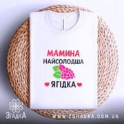 Футболка мамина найсолодша ягідка, біла, складена на круглій кошику, фон світло-сірий, принт з червоним текстом та малюнком ягід і листя.