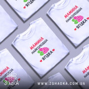 Футболка мамина найсолодша ягідка, біла, зображена на моделі з легким ракурсом, фон світло-фіолетовий, принт з червоним текстом та малюнком ягід і листя.