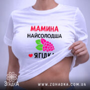 Футболка мамина найсолодша ягідка, біла, складена на поверхні, фон світло-сірий, принт з червоним текстом та малюнком ягід і листя, з етикеткою на комірці.