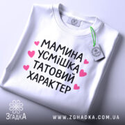 Футболка мамина усмішка, біла, складена на столі, фон світлий, принт чорний з рожевими сердечками, текст розташований по центру, видно етикетку з зеленим шнурком.