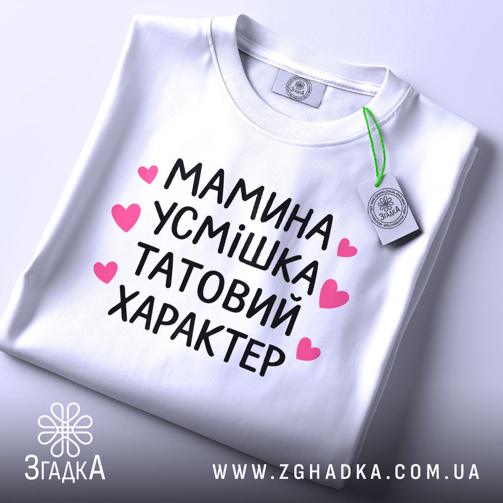 Футболка мамина усмішка, біла, складена на столі, фон світлий, принт чорний з рожевими сердечками, текст розташований по центру, видно етикетку з зеленим шнурком.