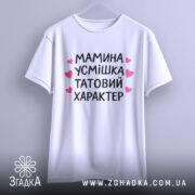 Футболка мамина усмішка, біла, підвішена на вішалці, фон світло-сірий, принт чорний з рожевими сердечками, текст розташований по центру, виглядає стильно.