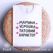 Футболка мамина усмішка, біла, складена на плетеній кошику, фон світлий, принт чорний з рожевими сердечками, текст розташований по центру, видно круглу горловину.