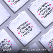 Футболка мамина усмішка, біла, на моделі, з легким ракурсом спереду, фон світло-фіолетовий, принт чорний з рожевими сердечками, текст читається чітко.