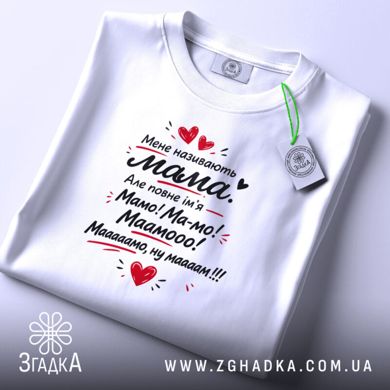 Футболка мене називають мама. На фото представлена біла футболка, з верхнього ракурсу, на фоні світло-сірого кольору. Принт на футболці містить той же текст, що й попередня, з червоними сердечками, а також видно етикетку з зеленим шнурком.