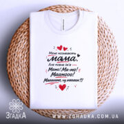 Футболка мене називають мама. На фото зображена біла футболка, з верхнього ракурсу, на фоні плетеного кошика. Принт на футболці аналогічний, з текстом та сердечками, розташований на рівній поверхні.