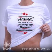 Футболка мене називають мама. На фото представлена біла футболка, з верхнього ракурсу, на фоні світло-сірого кольору. Принт на футболці містить той же текст, що й попередня, з червоними сердечками, а також видно етикетку з зеленим шнурком.