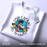 Футболка мій гараж мої правила, складена біла футболка на світлому фоні, зображення принта з вовком в окулярах, який тримає ключі, текст чорного кольору, етикетка з зеленим шнурком.