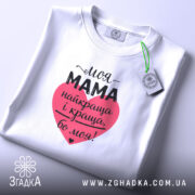 Футболка моя мама білого кольору, складена на поверхні, фон світло-сірий, принт у формі серця червоного кольору з текстом чорного кольору, на етикетці зелена стрічка.