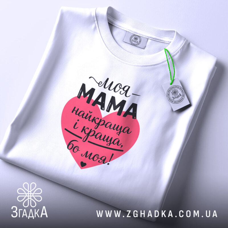 Футболка моя мама білого кольору, складена на поверхні, фон світло-сірий, принт у формі серця червоного кольору з текстом чорного кольору, на етикетці зелена стрічка.