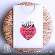 Футболка моя мама білого кольору, складена на кошику з ротанга, фон світло-сірий, принт у формі серця червоного кольору з текстом чорного кольору.