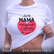 Футболка моя мама білого кольору, складена на поверхні, фон світло-сірий, принт у формі серця червоного кольору з текстом чорного кольору, на етикетці зелена стрічка.